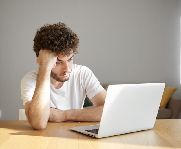 Horizontal shot of frustrated young unshaven male freelancer in white t-shirt using electronic device for distant work, having tired or bored look while working on urgent project early in the morning